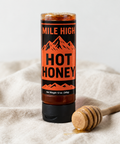 A squeeze bottle of Mile High Hot Honey topped with amber honey, standing on a beige linen cloth next to a wooden honey dipper.